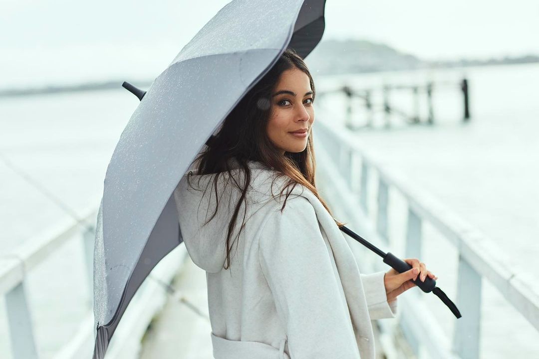 Young woman in a white coat stands under an umbrella on a wooden pier, looking over her shoulder with a slight smile on her face.