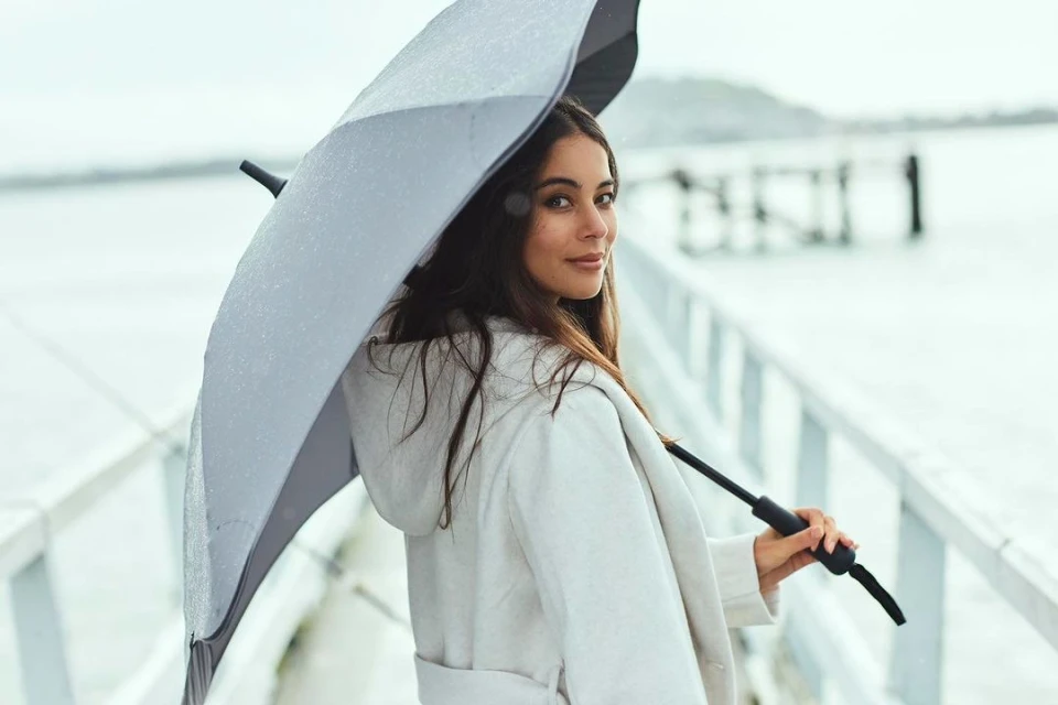 Young woman in a white coat stands under an umbrella on a wooden pier, looking over her shoulder with a slight smile on her face.