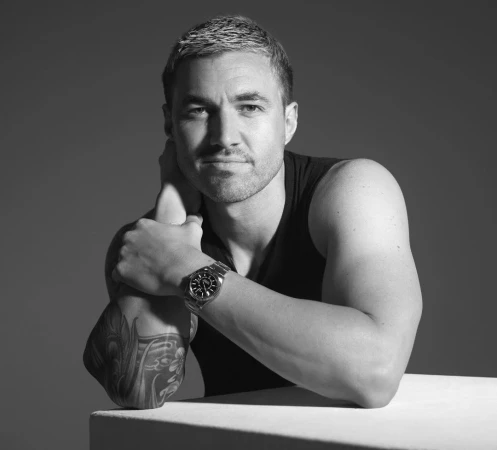 Portrait of a man in a dark tank top with a silver watch and arm tattoo resting on a white surface against a grey background.