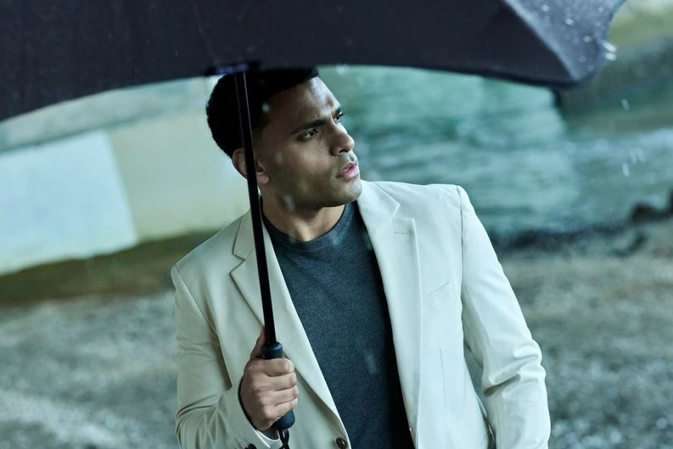 Man in a cream suit holding a black umbrella in the rain. He is facing to the right, looking up. He wears a gray shirt.