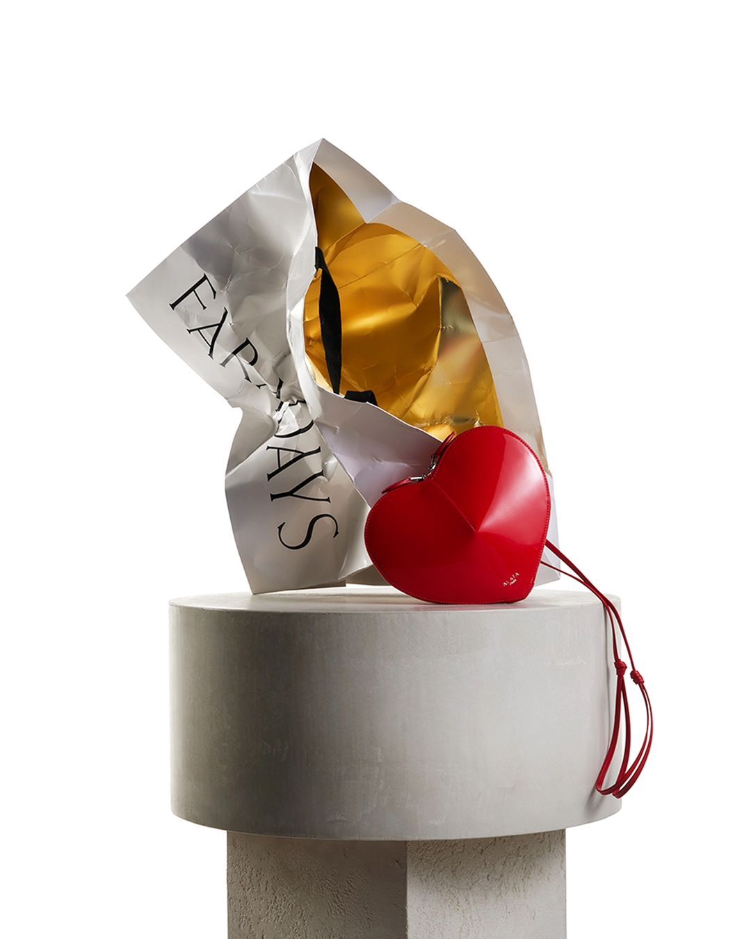 Red heart-shaped purse rests near a white paper shopping bag on a tiered concrete pedestal, isolated on a white studio background.
