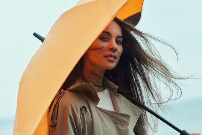 Woman holding a bright yellow umbrella wearing a beige trench coat against an overcast sky