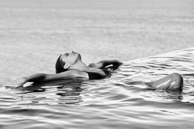 Black and white photograph of a woman floating in an infinity pool with her arms outstretched.