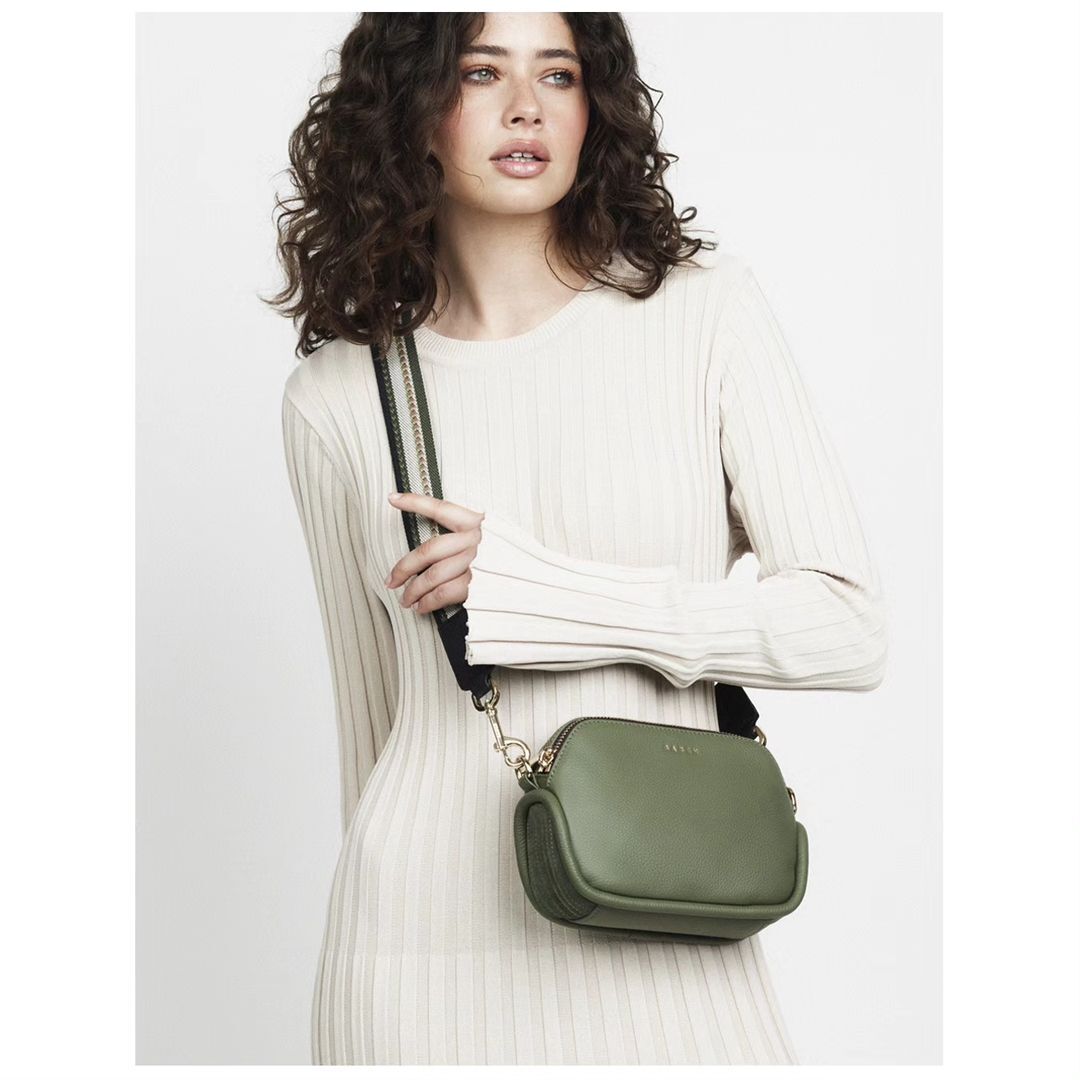 Woman wearing a light colored ribbed dress and holding a small olive green cross body bag with a black shoulder strap.