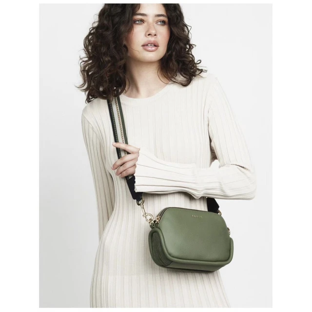 Woman wearing a light colored ribbed dress and holding a small olive green cross body bag with a black shoulder strap.