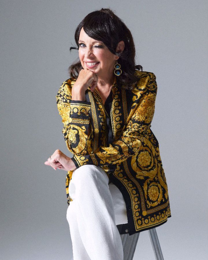 Adult woman seated on a step stool with a winking expression, wearing a patterned gold and black jacket and white pants in a studio.