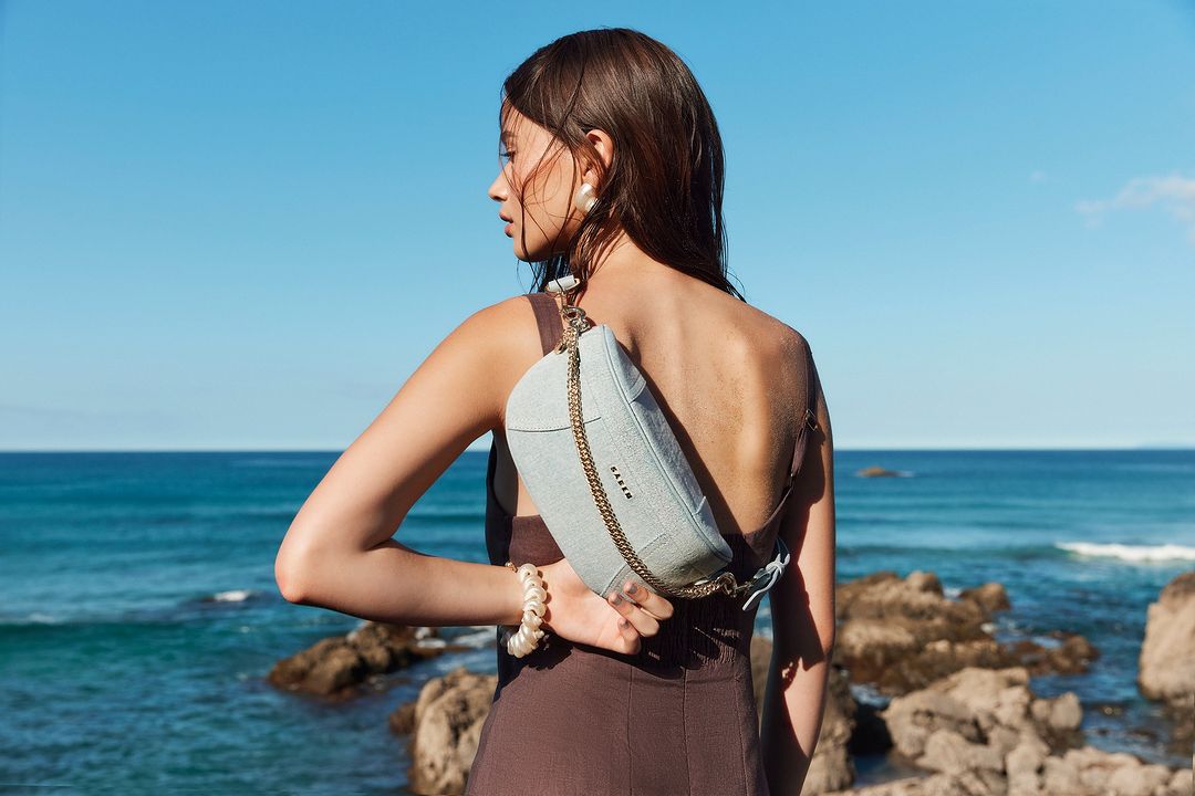 Woman in brown dress with purse stands near blue ocean and rocky coastline on sunny day in fashion editorial outdoor lifestyle photo.