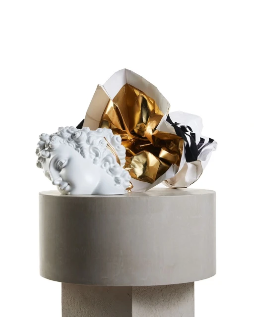 Modern still life featuring a white sculpture head on a plinth beside gold wrapping paper, set against a minimalist, isolated white backdrop.