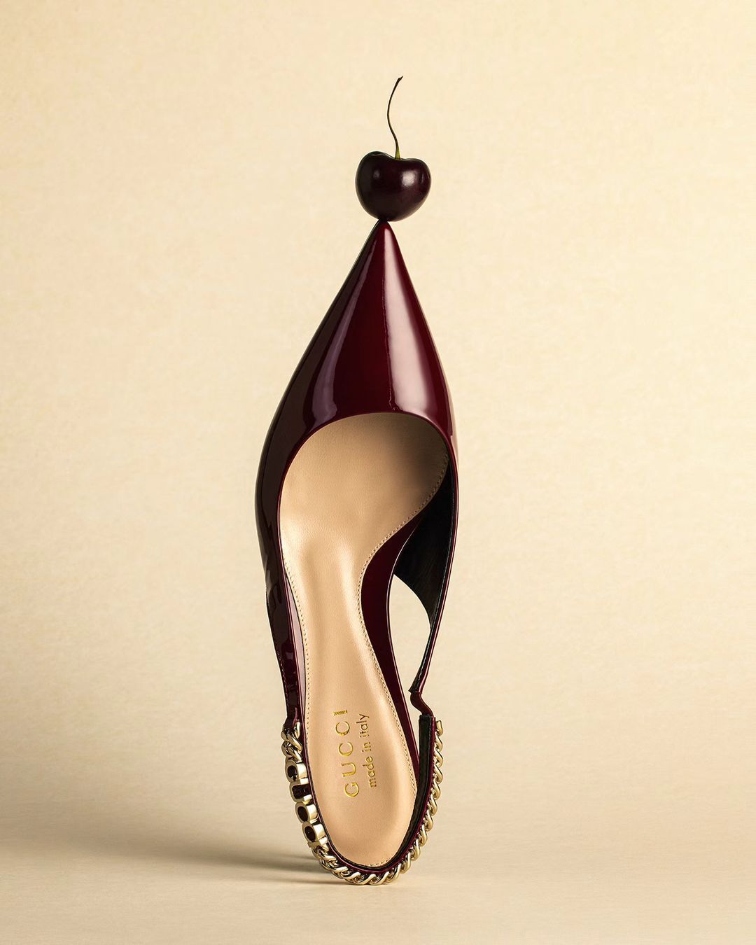 High-heeled slingback pump with a burgundy patent leather upper, gold-tone chain trim, and cherry on the toe against a cream background.