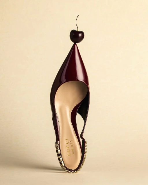 High-heeled slingback pump with a burgundy patent leather upper, gold-tone chain trim, and cherry on the toe against a cream background.