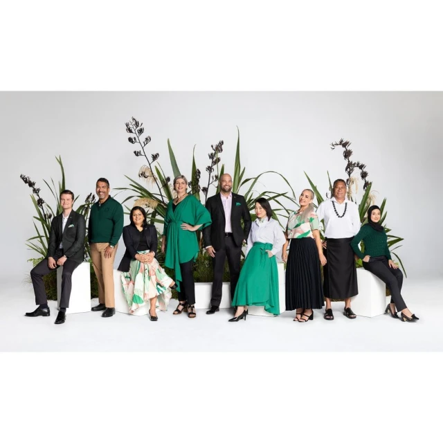Group portrait of nine diverse adults in green-accented business attire, posing in a photo studio with plant decorations against a white background.