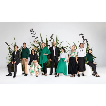 Group portrait of nine diverse adults in green-accented business attire, posing in a photo studio with plant decorations against a white background.