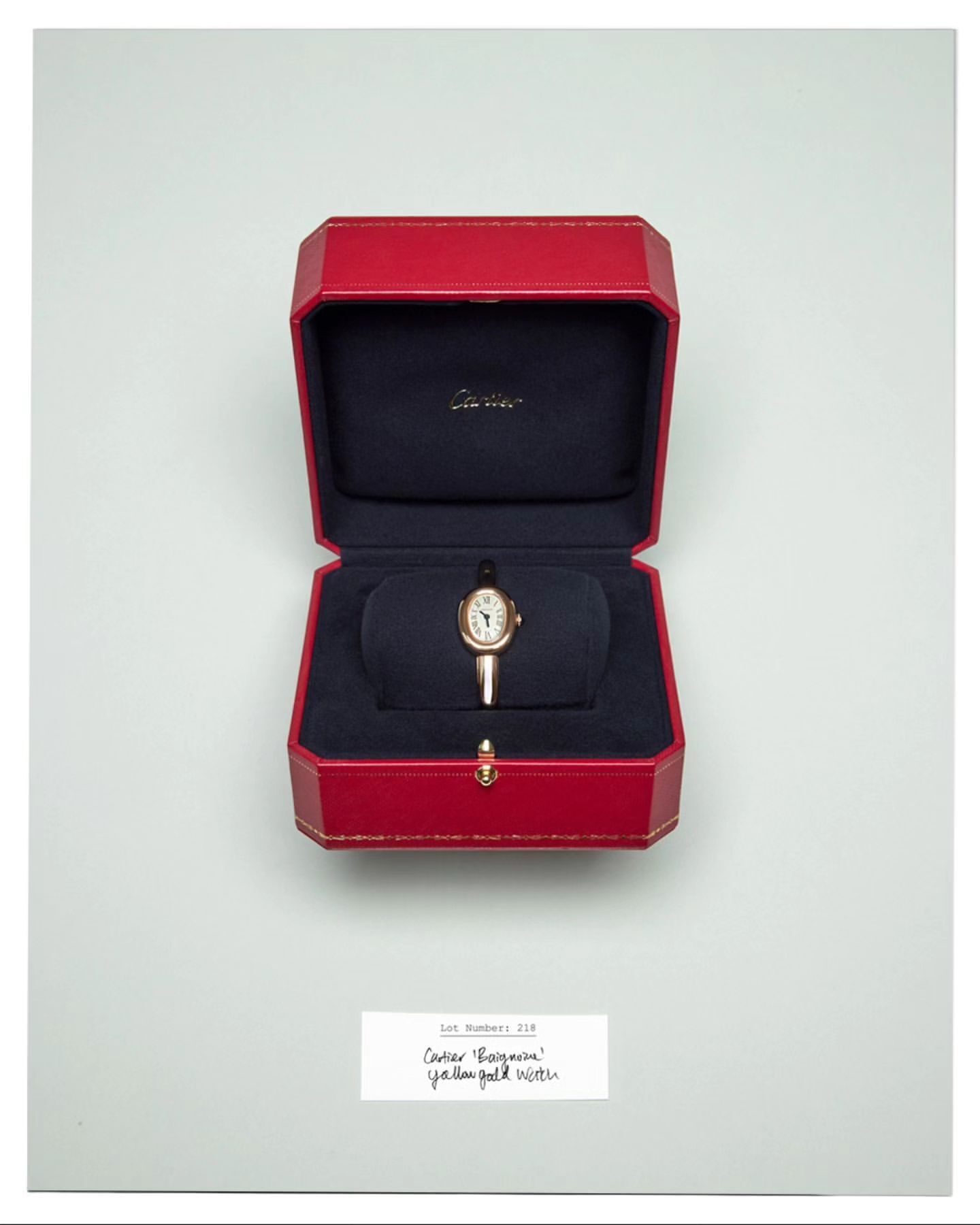 Close-up of a gold wristwatch displayed in its original red box with navy blue lining on a white background. Watch faces up.