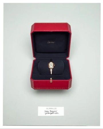Close-up of a gold wristwatch displayed in its original red box with navy blue lining on a white background. Watch faces up.