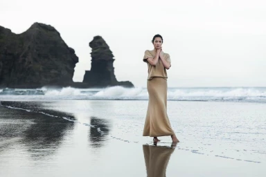 Woman in a beige knit dress standing on a wet sandy beach near rocky outcrops with ocean waves.