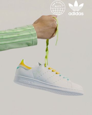 Adidas Stan Smith sneaker held up by a hand with green shoelaces against a neutral background.  Features Adidas logo and End Plastic Waste emblem.