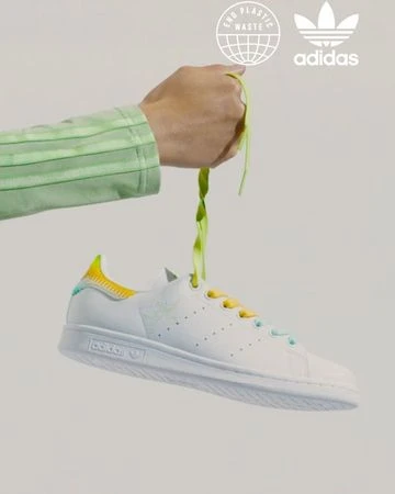 Adidas Stan Smith sneaker held up by a hand with green shoelaces against a neutral background.  Features Adidas logo and End Plastic Waste emblem.