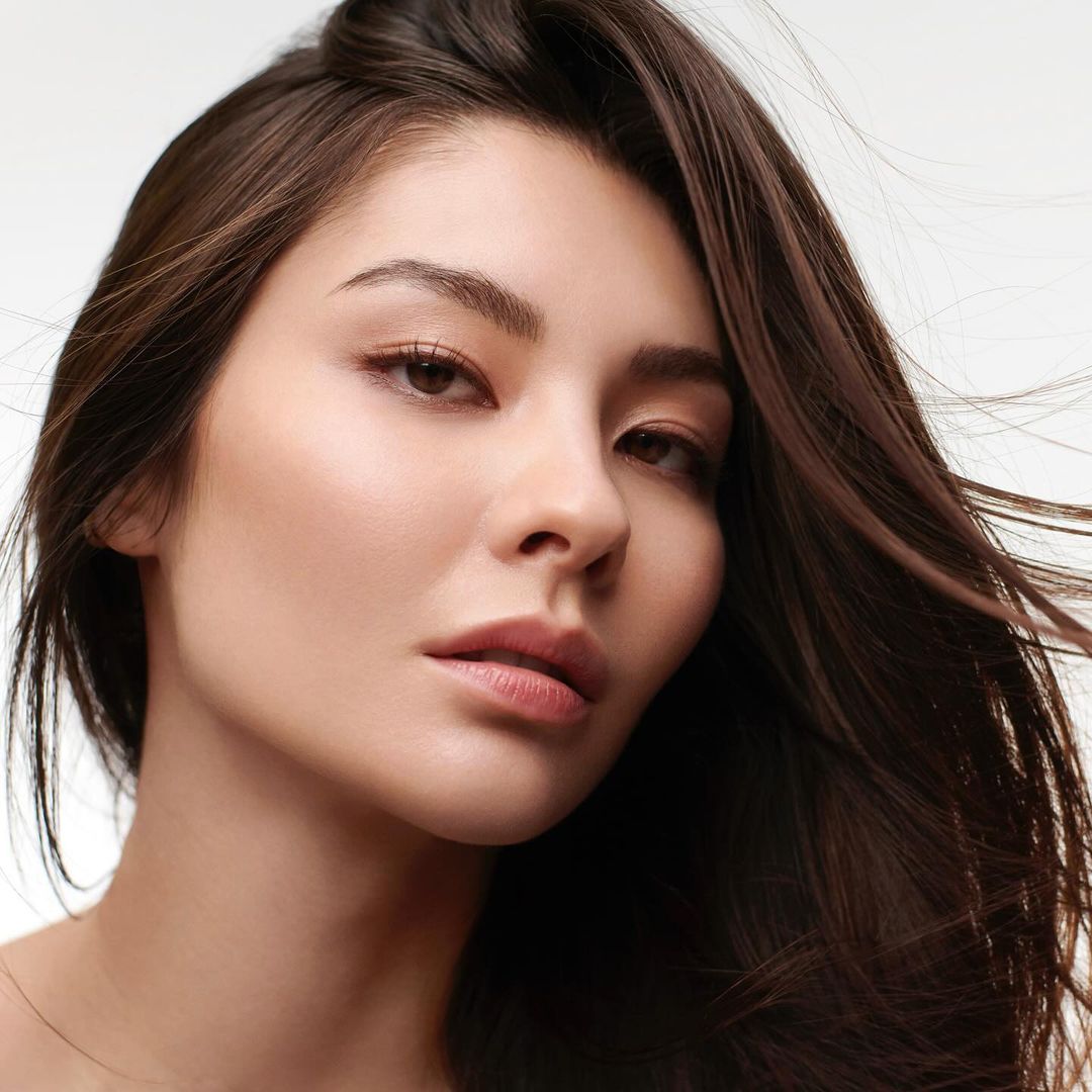 Portrait of a young adult woman with long brown hair and neutral facial expression against a clean white background in eye-level close-up.