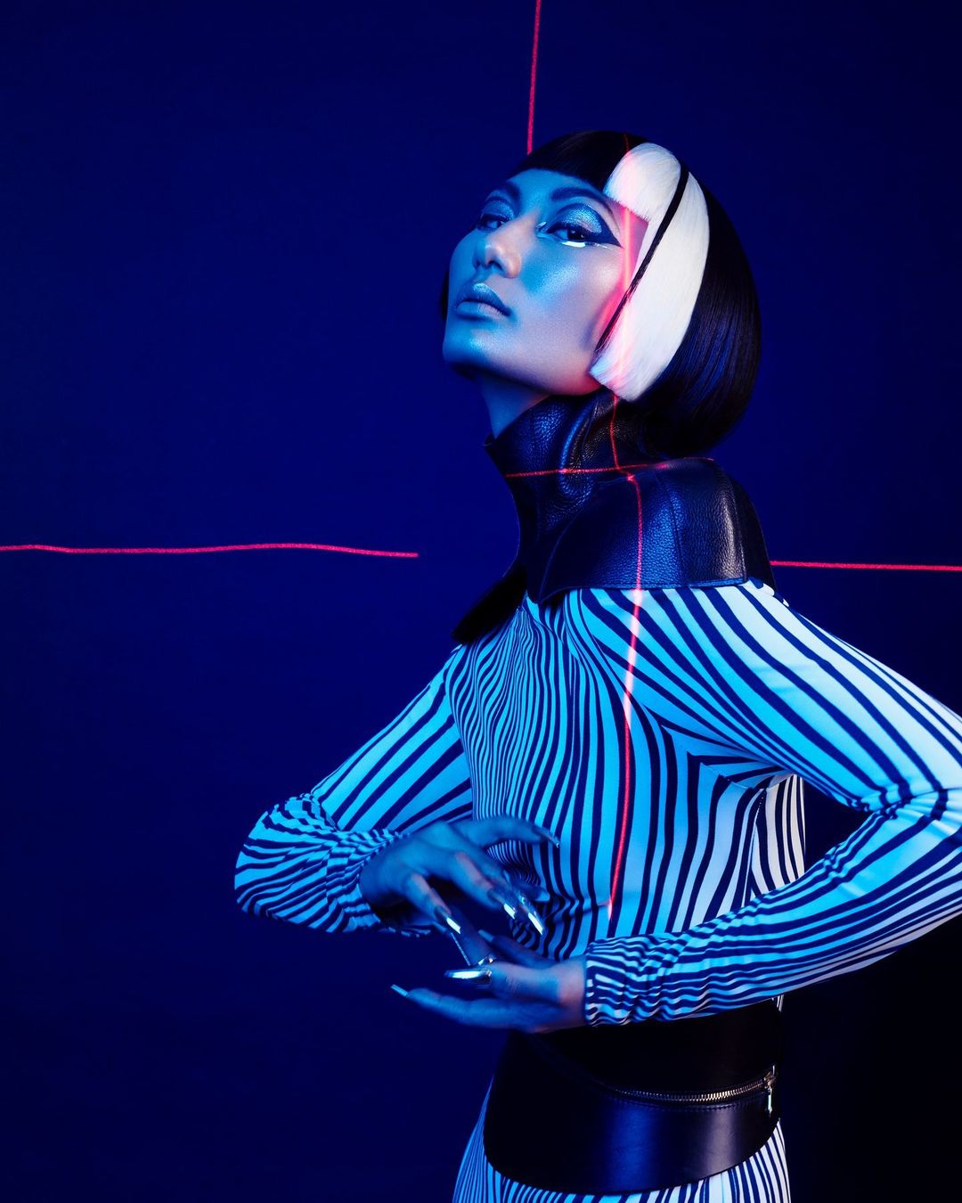 Woman with graphic makeup and striped dress poses in front of dark blue background with pink laser lines. Striking and modern portrait.