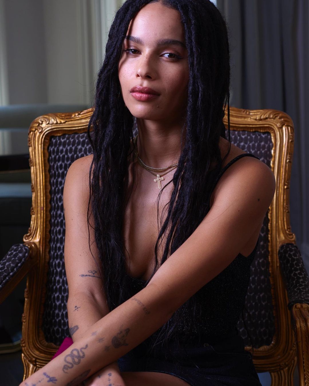 Portrait of Zoe Kravitz with dark hair, wearing a dark dress and gold necklaces, seated in an ornate gold chair.