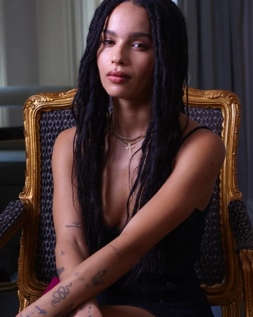 Portrait of Zoe Kravitz with dark hair, wearing a dark dress and gold necklaces, seated in an ornate gold chair.