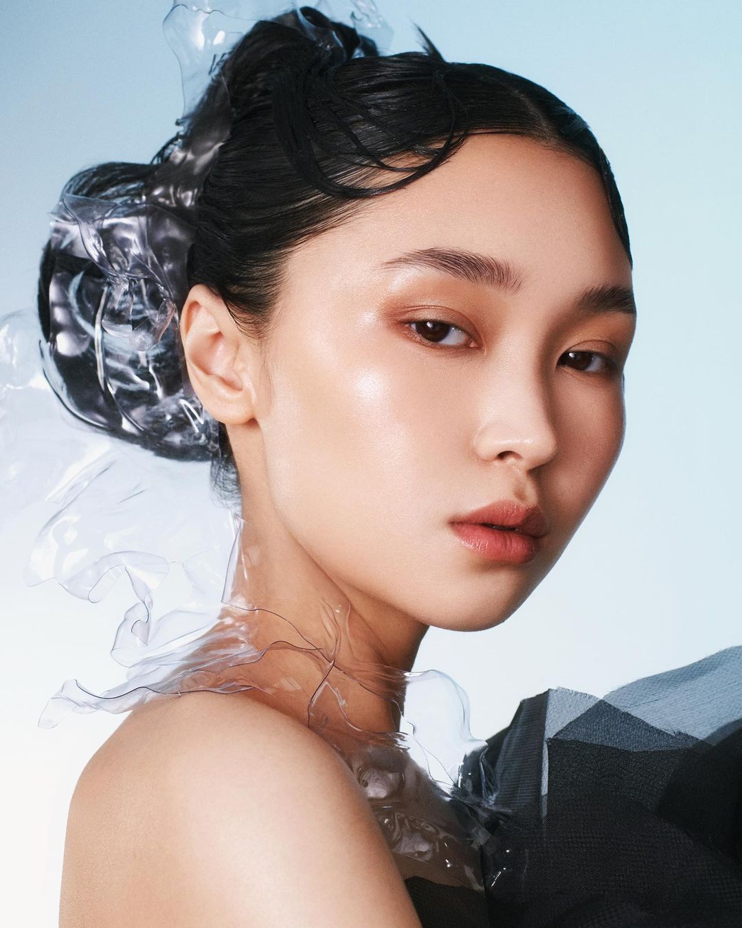 Close-up beauty portrait of a young Asian woman with slicked-back hair and clear plastic accents against a soft blue background.