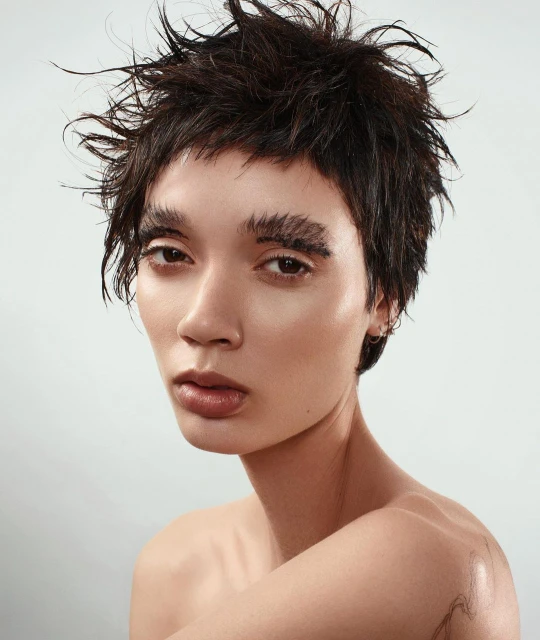 Close-up portrait of a young adult woman with short, spiky dark hair and dramatic makeup against a pale background, beauty shot.