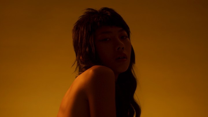 Portrait of an Asian woman with dark hair looking back over her shoulder, bathed in a warm yellow glow against a yellow background.