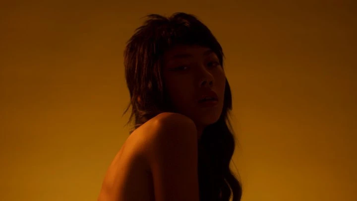 Portrait of an Asian woman with dark hair looking back over her shoulder, bathed in a warm yellow glow against a yellow background.