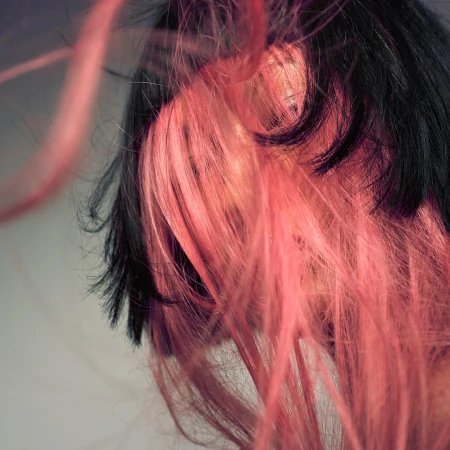 Close-up portrait of a person with dark and pink dyed hair blowing across their face against a neutral background.