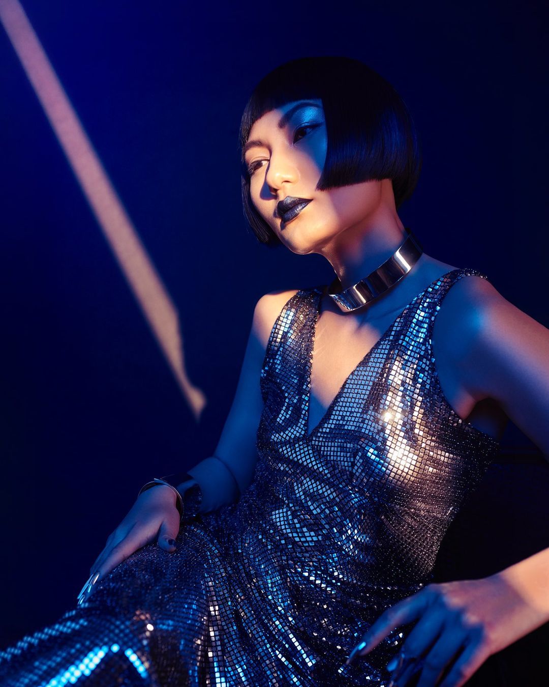 Glamorous Asian woman poses in sequin dress and bold makeup under blue light, showcasing fashion and beauty in a studio setting.