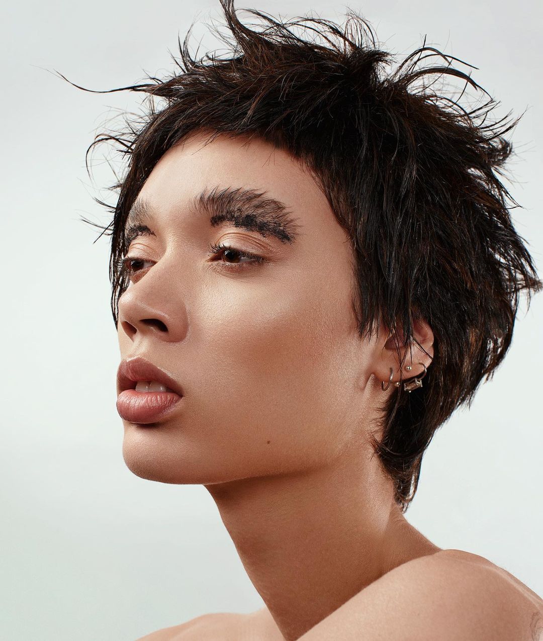Close-up studio portrait of a young adult with short dark hair, light skin, light pink lips, and dramatic feathered eyebrows looking left.