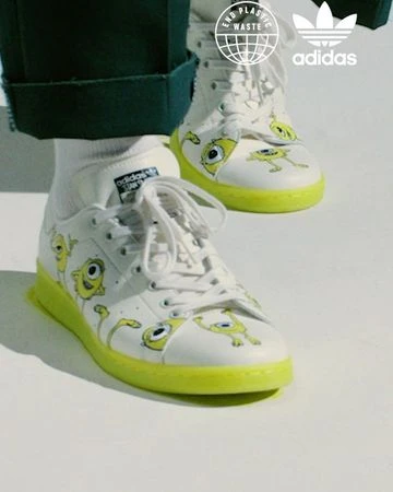 Close-up of white Adidas Stan Smith sneakers with cartoon monster graphics and neon yellow soles, displaying the Adidas logo and End Plastic Waste symbol.