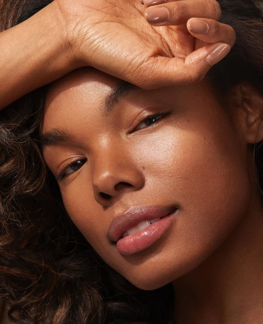 Close-up portrait of a young adult woman with tan skin, brown curly hair, and natural makeup, posing with her hand near her forehead.