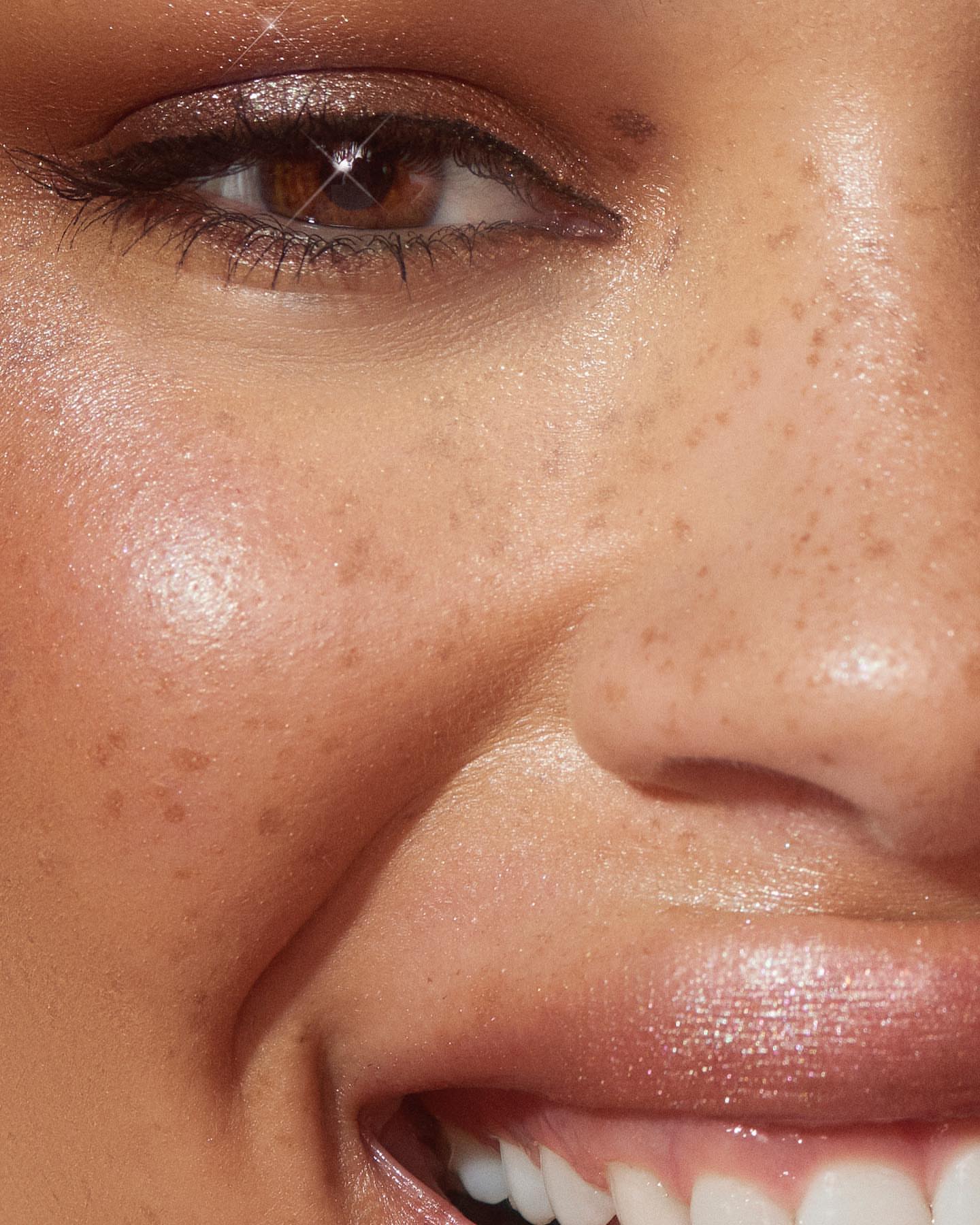 Close-up of a smiling adult woman with brown eyes, freckled skin, lip gloss, and cosmetic eye makeup, captured in a beauty portrait.
