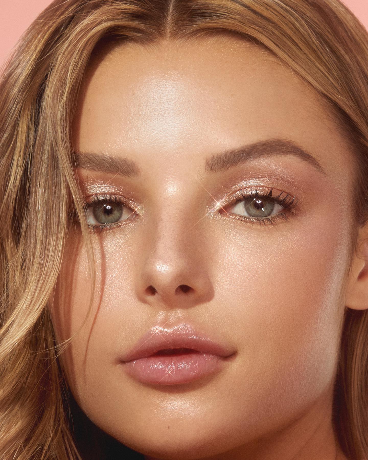 Close-up portrait of a young woman with blonde hair, fair skin, and light eyes, showcasing a natural makeup look on a soft pink background.