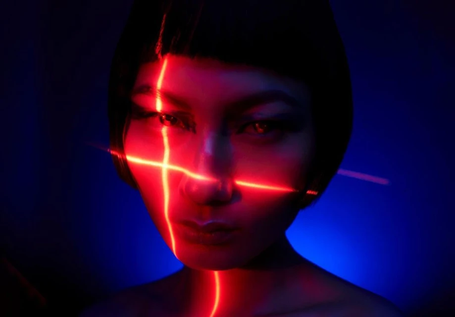 Close-up portrait of an Asian woman with short black hair and red laser lights crossing her face, set against a dark blue backdrop.