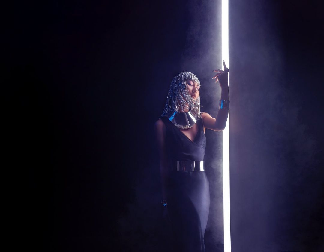 Woman in dark dress and silver headdress posing next to a vertical light tube in a dark studio with hazy atmosphere.