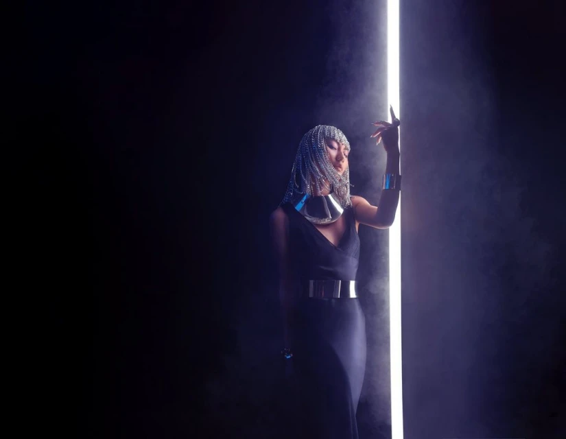 Woman in dark dress and silver headdress posing next to a vertical light tube in a dark studio with hazy atmosphere.