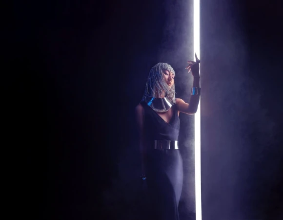 Woman in dark dress and silver headdress posing next to a vertical light tube in a dark studio with hazy atmosphere.