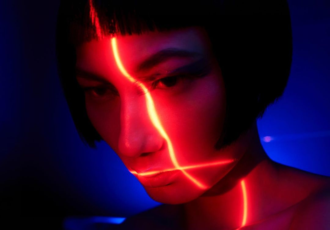 Portrait of a woman with short dark hair and bright red geometric lighting patterns on her face against a deep blue background.