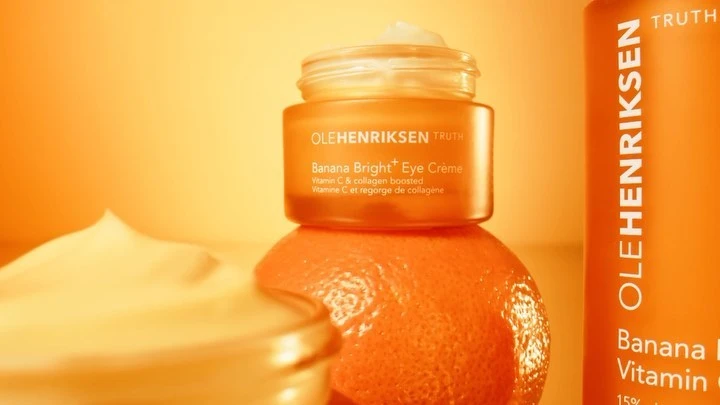 Olehenriksen Banana Bright Eye Cream jar and product bottle in a still life, with an orange and a cream-colored smear.