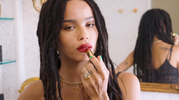 Adult woman with dreadlocks applying red lipstick in front of a mirror; she's wearing jewelry and a black top in an indoor environment.