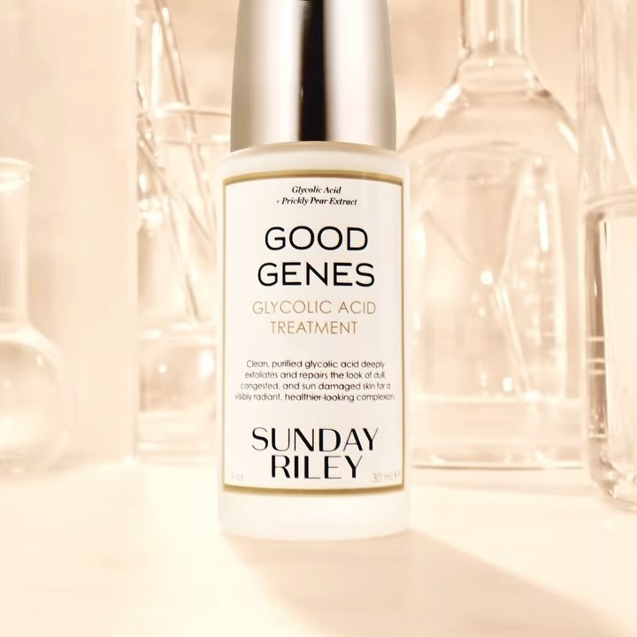 Close-up of Sunday Riley Good Genes Glycolic Acid Treatment bottle with clear lab glassware in background, pale yellow tones.