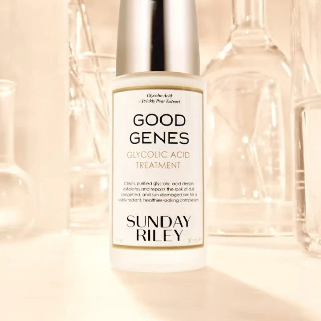 Close-up of Sunday Riley Good Genes Glycolic Acid Treatment bottle with clear lab glassware in background, pale yellow tones.