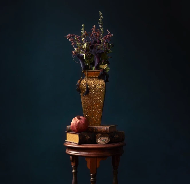 Still life arrangement featuring a gold vase with dried flowers, books, a pomegranate, and an eye charm on a dark teal background.