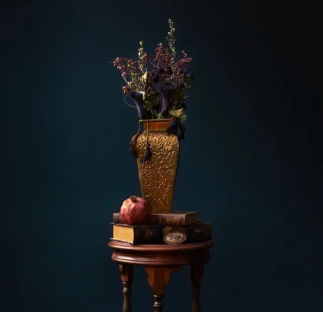 Still life arrangement featuring a gold vase with dried flowers, books, a pomegranate, and an eye charm on a dark teal background.