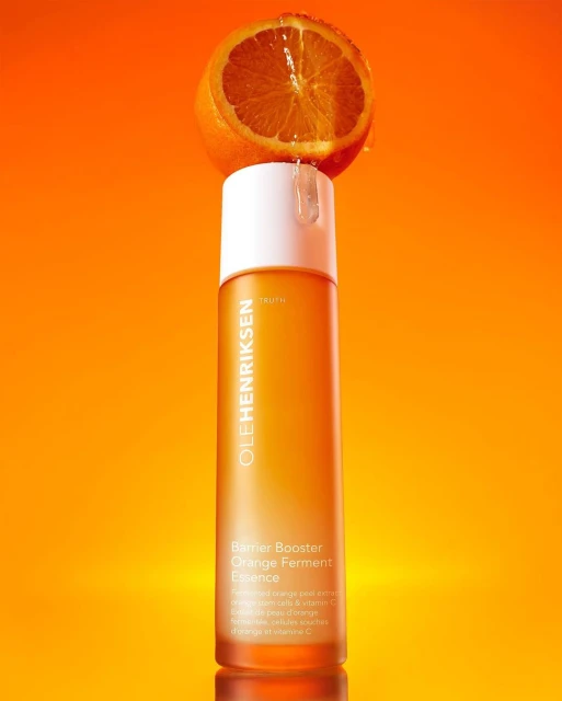 Ole Henriksen Barrier Booster Orange Ferment Essence bottle with half orange on top against an orange gradient background.
