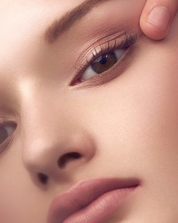 Close-up studio shot of a fair-skinned adult female face with light brown hair, featuring makeup, with a finger pulling on the upper eyelid.