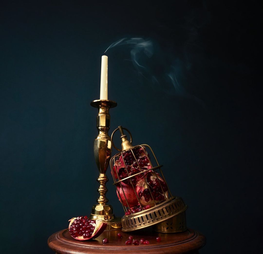 Still life photograph of a pomegranate in a cage, extinguished candle on a brass candlestick, and pomegranate seeds on a wooden table.